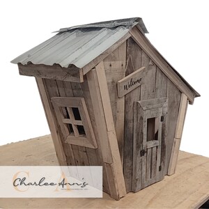 May include: A wooden miniature house with a gray metal roof, a small window, and a door with a sign that says "Welcome". The house is tilted to the side and is made of reclaimed wood.