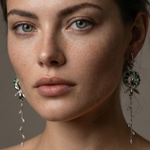 Puede incluir: Primer plano de un rostro con piel clara, pecas y ojos gris-azules. La persona lleva pendientes largos y colgantes de plata con detalles de piedras preciosas verdes. Los pendientes presentan un diseño de corona y un delicado tallo con hojas.