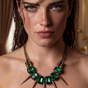 Pode incluir: Close-up de uma mulher usando um colar marcante. O colar apresenta grandes pedras retangulares verde-esmeralda e pontas douradas. A mulher tem cabelo escuro e molhado e está usando um top marrom.