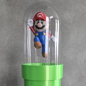 May include: A decorative display featuring a Mario figurine inside a clear dome. The figurine is atop a green pipe structure, which is set on a brown, brick-like base. The Mario figure is wearing his signature red hat, blue overalls, and white gloves.