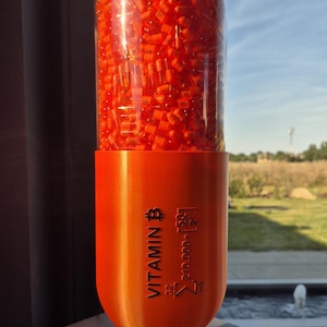 May include: A large orange capsule filled with smaller orange capsules. The capsule is labeled "VITAMIN B" and has a mathematical formula printed on the side.  The capsule is sitting on a black base.