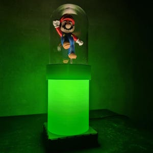 May include: A green-glowing lamp featuring a Super Mario figurine inside a glass dome. The lamp has a cylindrical base and a square base. The Mario figure is in his signature red and blue outfit, with his arm raised in a wave.