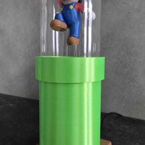 May include: A decorative lamp featuring a Mario figurine inside a clear glass dome. The figurine is atop a bright green pipe, which sits on a brown brick-like base. The lamp is a unique piece of home decor.