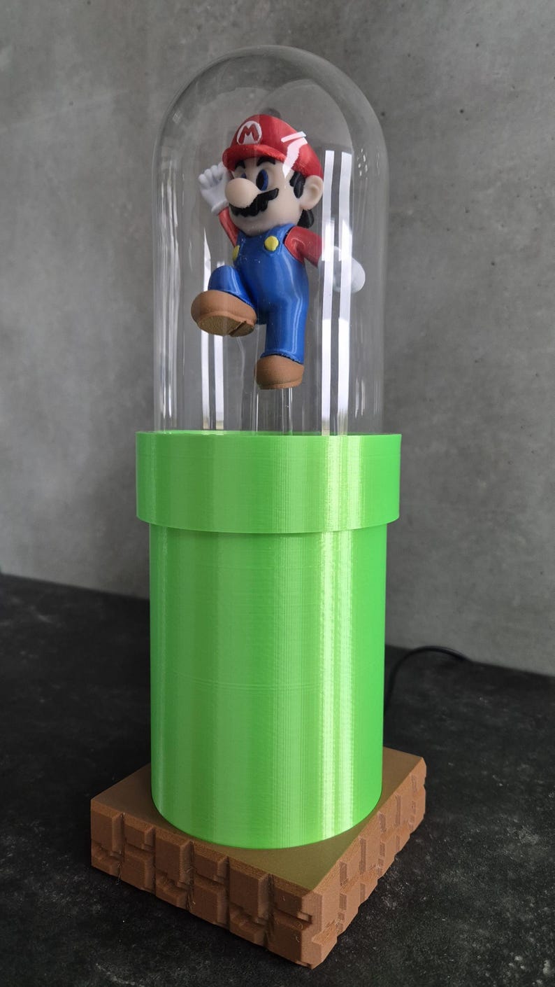 May include: A decorative lamp featuring a Mario figurine inside a clear glass dome. The figurine is atop a bright green pipe, which sits on a brown brick-like base. The lamp is a unique piece of home decor.