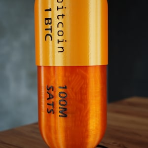 Puede incluir: Un objeto de forma de cápsula naranja impreso en 3D con el texto "bitcoin 1 BTC 100M SATs" impreso en él. El objeto está sentado sobre una base plateada.
