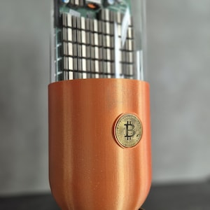 Könnte beinhalten: Eine transparente Glaskapsel, die eine kleine Platine mit einem Kühlventilator enthält. Die Kapsel ist an einer kupferfarbenen Basis befestigt, an der sich seitlich eine goldfarbene Bitcoin-Münze befindet.