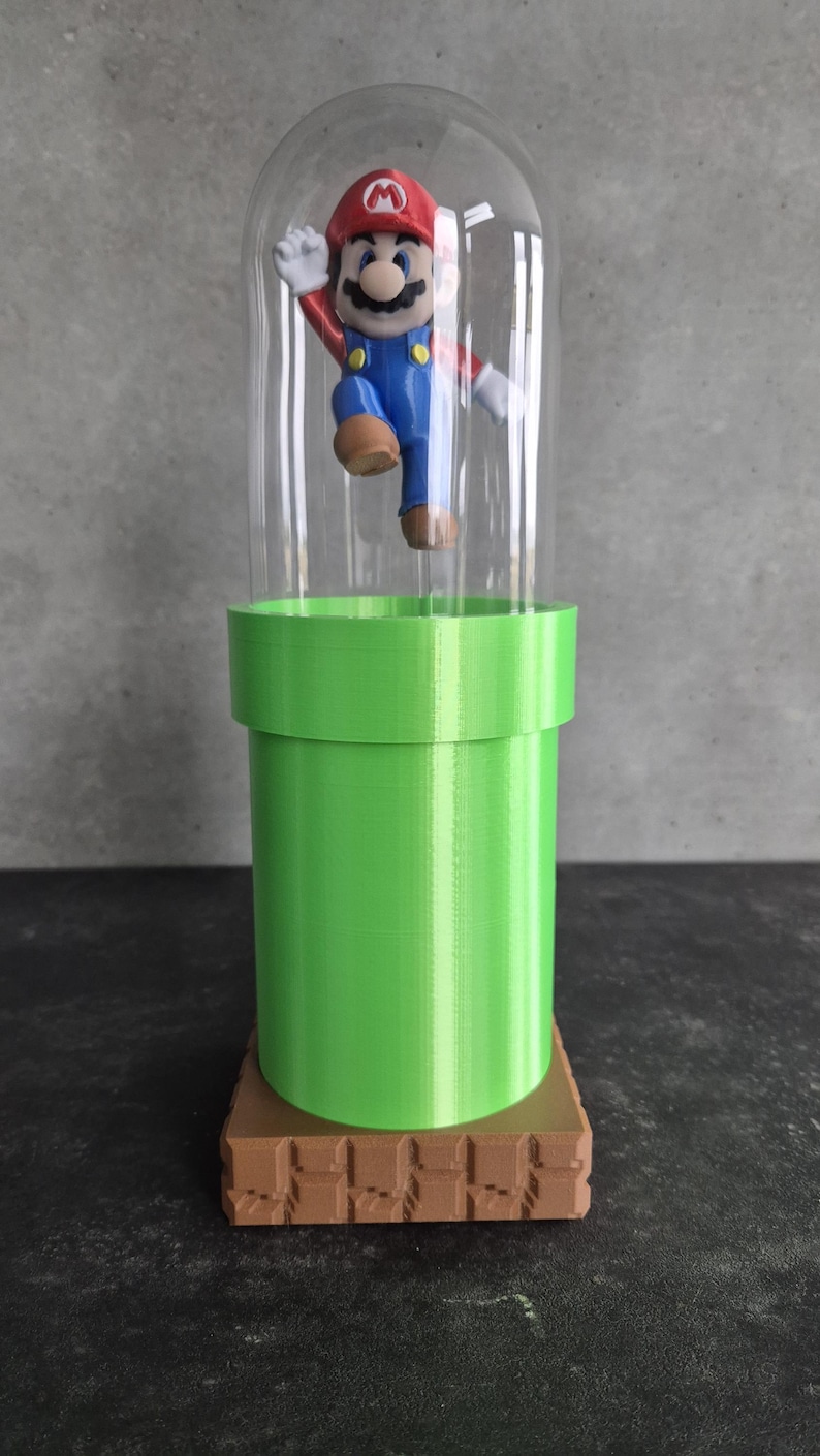 May include: A decorative display featuring a Mario figurine inside a clear dome. The figurine is atop a green pipe structure, which is set on a brown, brick-like base. The Mario figure is wearing his signature red hat, blue overalls, and white gloves.