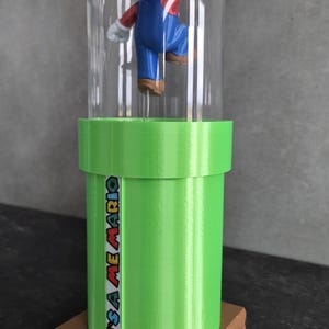 May include: A 3D-printed figurine of Mario inside a clear dome, atop a green pipe with the text "IT'S A ME MARIO". The base is brown, and the figure is in a jumping pose, wearing his signature red hat and blue overalls.
