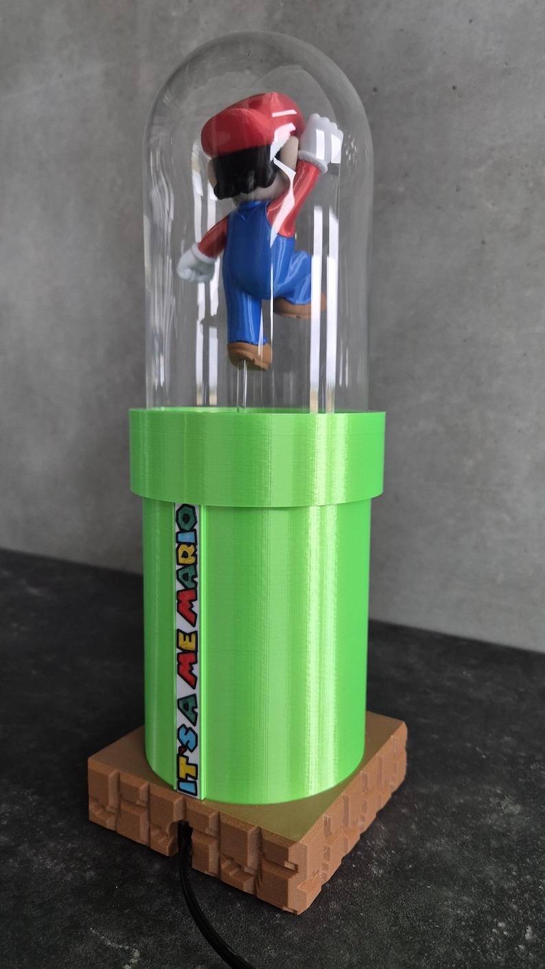 May include: A 3D-printed figurine of Mario inside a clear dome, atop a green pipe with the text "IT'S A ME MARIO". The base is brown, and the figure is in a jumping pose, wearing his signature red hat and blue overalls.