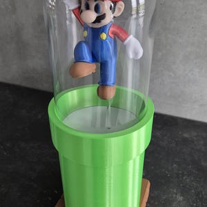May include: A 3D-printed figurine of Mario, wearing a red cap and blue overalls, inside a clear glass dome. The dome sits atop a green pipe, which is on a brown, textured base. The figurine is in a jumping pose.