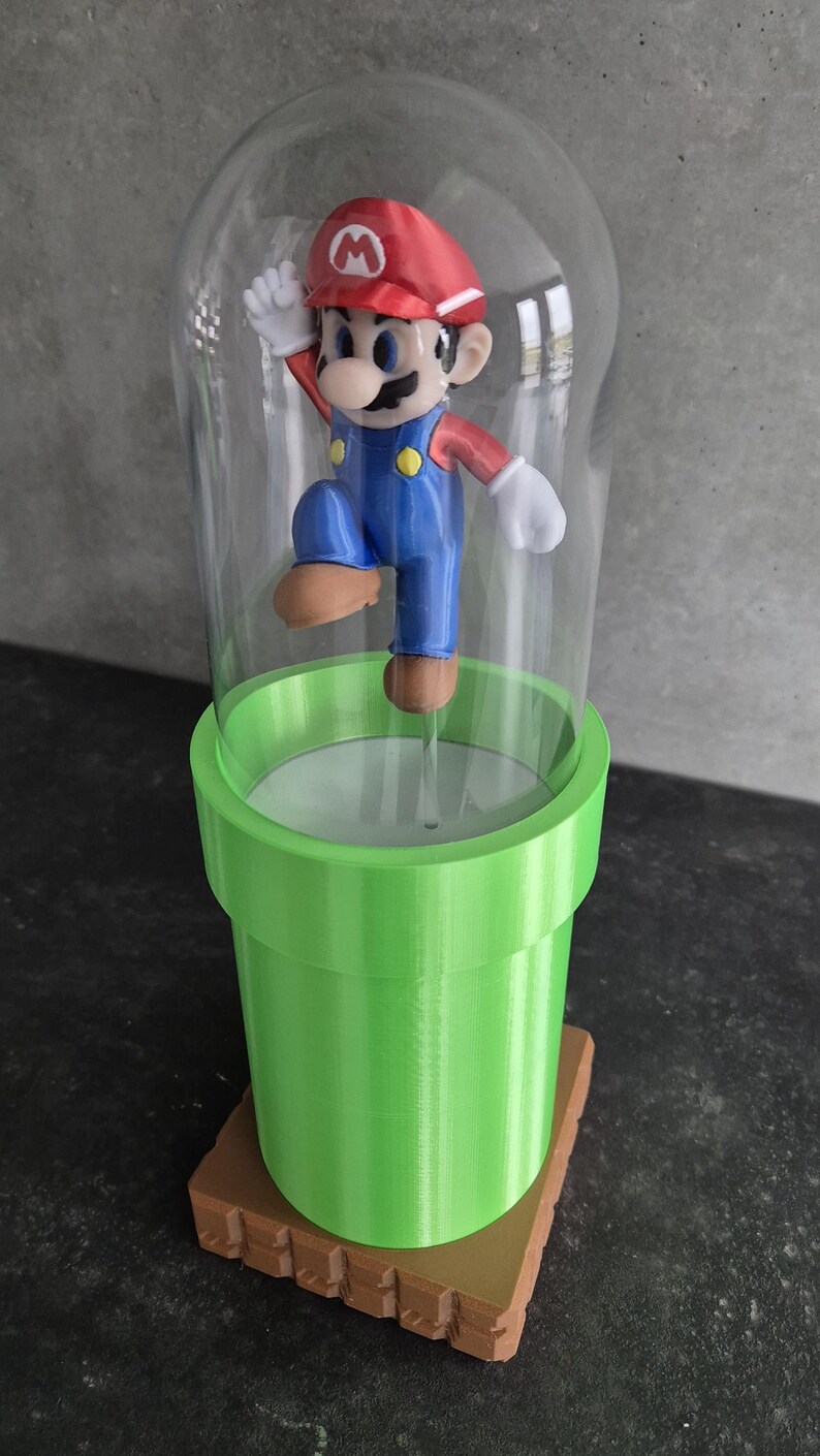 May include: A 3D-printed figurine of Mario, wearing a red cap and blue overalls, inside a clear glass dome. The dome sits atop a green pipe, which is on a brown, textured base. The figurine is in a jumping pose.