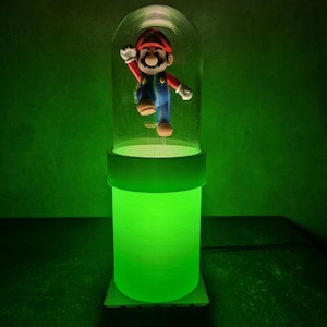 May include: A green-glowing lamp featuring a figurine of Mario inside a clear dome. The lamp has a cylindrical base and a circular top, with the Mario figure in a jumping pose. The background is a solid green.