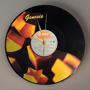 May include: A round clock made from a black vinyl record. The clock face features the Genesis logo in yellow, with a design of orange and yellow geometric shapes. The clock hands are gold-colored.
