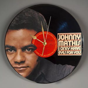 May include: A circular clock crafted from a black vinyl record. The clock face displays a portrait of Johnny Mathis and a red record label. The text "JOHNNY MATHIS ONLY HAVE EYES FOR YOU" is visible. Gold-coloured clock hands.