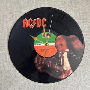 Könnte beinhalten: Eine schwarze Vinyl-Schallplattenuhr mit dem AC/DC-Logo in Rot. Das Zifferblatt zeigt das Albumetikett mit Songtiteln und einem Bandbild. Goldfarbene Zeiger zeigen die Uhrzeit an. Ein einzigartiger Dekorationsartikel.