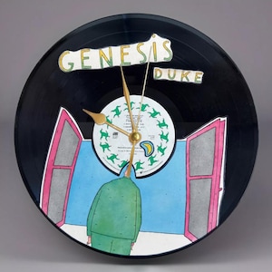 May include: A black vinyl record clock with the words "GENESIS DUKE" in green and yellow. The clock face features a whimsical illustration of a person in a green suit looking out a window. Gold clock hands.