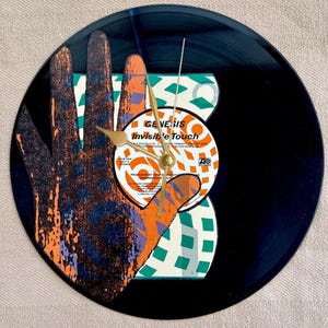 May include: A round clock made from a black vinyl record. The clock face features a hand design with orange, purple, and white accents. The center of the clock displays the album art for "Invisible Touch" by Genesis. Gold-colored clock hands.