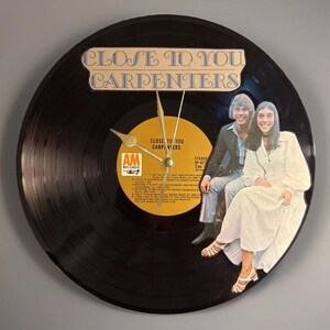 Puede incluir: Un reloj de vinilo negro con el texto "CLOSE TO YOU CARPENTERS" y una foto de dos personas. La esfera del reloj presenta la etiqueta del álbum y manecillas doradas.