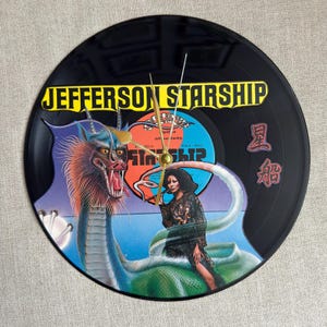 May include: A black vinyl record clock featuring the band name "Jefferson Starship" in yellow lettering. The clock face displays a colorful illustration of a dragon and a woman. Gold clock hands indicate the time.