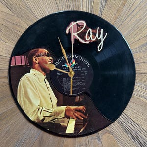 May include: A black vinyl record clock featuring a photo of Ray Charles playing the piano. The clock has gold-colored hands and the name "Ray" in a cursive font. The record label is visible.