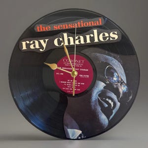 May include: A black vinyl record clock featuring the text "the sensational ray charles". The clock face includes a portrait of Ray Charles and a red label with song titles. Gold-colored clock hands indicate the time.