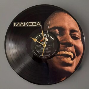 Peut inclure: Une horloge en vinyle noir avec un portrait d'une femme souriante. Le cadran de l'horloge comprend le texte "MAKEBA" et une petite étiquette de disque. Les aiguilles dorées indiquent l'heure.