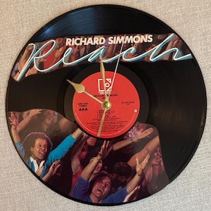 May include: A black vinyl record clock featuring the album art for Richard Simmons' "Reach." The clock face has a red center label with gold clock hands. The album art shows a crowd of people with their arms raised.