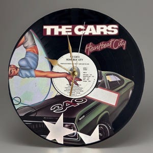 May include: A black vinyl record clock featuring the album art from The Cars' "Heartbeat City." The clock face includes the album title and a green car graphic. Gold-colored clock hands and a central clock mechanism are visible.