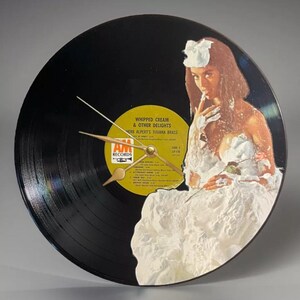 May include: A black vinyl record clock featuring a woman covered in whipped cream. The clock face includes the album title "Whipped Cream & Other Delights" by Herb Alpert's Tijuana Brass. Gold clock hands indicate the time.