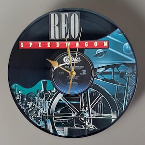 May include: A round wall clock made from a vinyl record. The clock face features the band name "REO Speedwagon" and album art with a train illustration. Gold-colored clock hands indicate the time.
