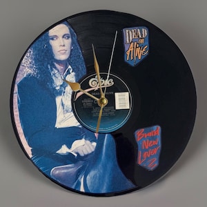 May include: A circular wall clock crafted from a black vinyl record featuring a portrait of a person with long dark hair, the text Dead or Alive, and the text Brand New Lover. The clock has gold-colored hands and a central Epic record label.