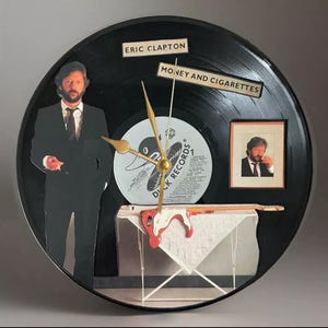 May include: A unique wall clock made from a black vinyl record. The clock features images of Eric Clapton, the text "Eric Clapton" and "Money and Cigarettes", and a small framed photo. Gold clock hands and a guitar are also included.