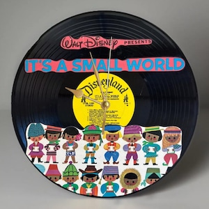 Könnte beinhalten: Eine schwarze Vinyl-Schallplattenuhr mit den Schriftzügen "It's a Small World" und "Disneyland". Das Zifferblatt zeigt eine farbenfrohe Illustration von Kindern in verschiedenen Outfits. Goldene Zeiger zeigen die Uhrzeit an.