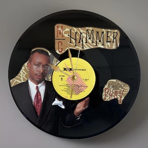 May include: A black vinyl record clock featuring a portrait of MC Hammer. The clock face includes the text "MC Hammer" and "Pray". The clock hands are gold-colored, and the center label reads "Burtamer".