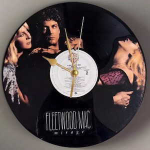 May include: A round wall clock made from a black vinyl record. The clock face features the Fleetwood Mac album cover "Mirage." Gold-colored hands indicate the time. The center label is white with black text. The album cover shows three people.
