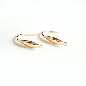 May include: A pair of gold-coloured earrings, each featuring a leaf-like shape with a curved hook and a small loop. The earrings are presented on a white backdrop, accentuating their polished finish. The design is simple and elegant.