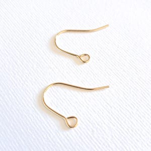 May include: Two gold-coloured earring hooks are shown against a white backdrop. The hooks have a curved design with a small loop at the bottom and a straight end. The hooks are made of a thin, shiny metal.
