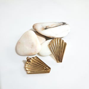 Pode incluir: Um par de brincos de metal dourado em forma de conchas em leque. Cada brinco apresenta uma série de estrias verticais que irradiam da base até à borda superior recortada. Um brinco mostra a face frontal, o outro mostra o verso.