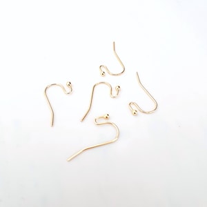Può includere: Diversi ganci per orecchini color oro sono sparsi su una superficie bianca. I ganci hanno un design curvo con una piccola sfera all'estremità. I ganci sono in metallo lucido.