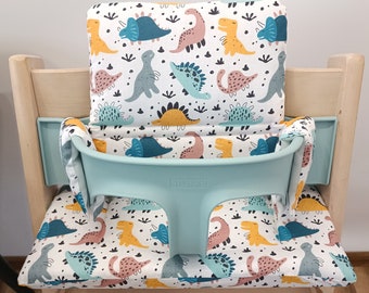 May include: A light blue high chair cushion with a white background and a dinosaur pattern. The cushion has a light blue strap and the text "STOKKE" is printed on the front.