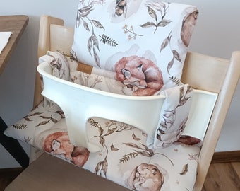 May include: A white high chair with a beige and white padded seat cover featuring a woodland animal print. The print includes foxes, squirrels, and leaves.