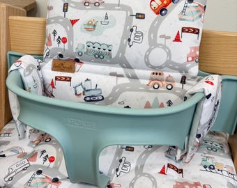 May include: A white high chair cushion with a gray and white road design featuring various vehicles, including a red fire truck, a yellow excavator, and a blue truck. The cushion is placed on a light blue high chair with the word "STOKKE" printed on the front.