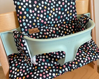 May include: A high chair cushion set featuring a black fabric with a multicolored polka dot pattern in white, pink, yellow, and teal. The set includes a backrest and seat pad, fitted with a light green plastic baby seat attachment labeled STOKKE.