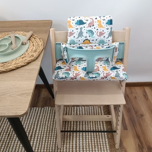 May include: A light wood high chair with a removable tray and a dinosaur-themed seat cushion. The cushion is white with colourful dinosaurs and green leaves.