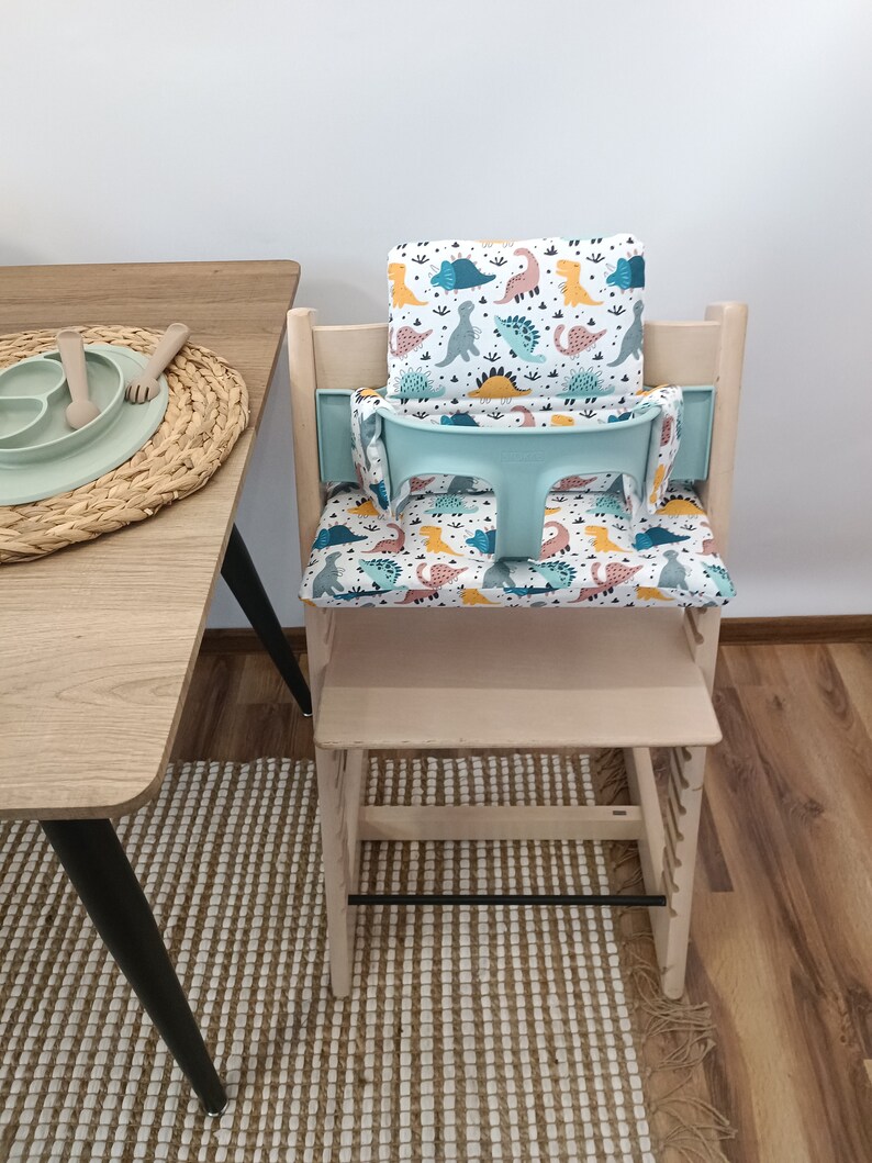 May include: A light wood high chair with a removable tray and a dinosaur-themed seat cushion. The cushion is white with colourful dinosaurs and green leaves.
