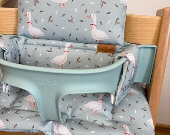 May include: A light blue high chair cushion with a white goose pattern and green leaves. The cushion has a built-in headrest and a strap to secure it to the chair. The text "Stokke" is printed on the cushion.