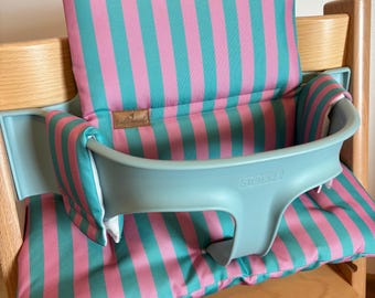 May include: A high chair cushion set featuring a vertical pink and teal striped pattern. The set includes a backrest pad and a seat pad, both fitted with a light blue plastic baby rail embossed with the text STOKKE. A small brown fabric label is attached.
