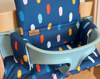 May include: A dark blue high chair cushion set featuring a pattern of vertical oval shapes in orange, yellow, light blue, and pink. The cushion includes a small brown rectangular label with the text trele morele and a light green plastic seat insert.