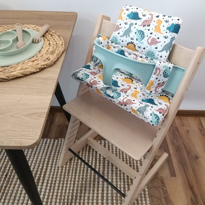 May include: A light wood high chair with a turquoise and white dinosaur print seat cushion and a matching back cushion. The chair is on a woven rug.