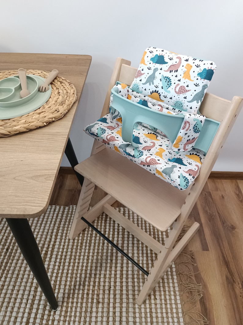 May include: A light wood high chair with a turquoise and white dinosaur print seat cushion and a matching back cushion. The chair is on a woven rug.